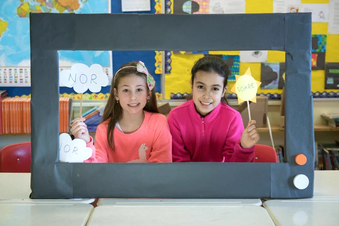 Two students role-played as weather anchors. The student on the left held two paper cut-outs of a cloud that reads, "nor." The student on the right held a paper cut-out of a sun that reads, "soare." 
