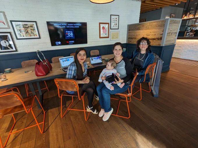 Three women and a baby sitting at a table going through a workshop but turned to the camera smiling to the picture.