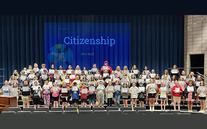 large group of students on risers in auditorium, large citizenship sign in background