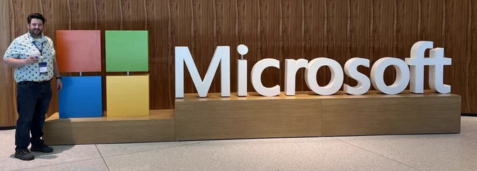 Sean in a blue shirt and blue jeans holding a coffee in front of a Microsoft sign