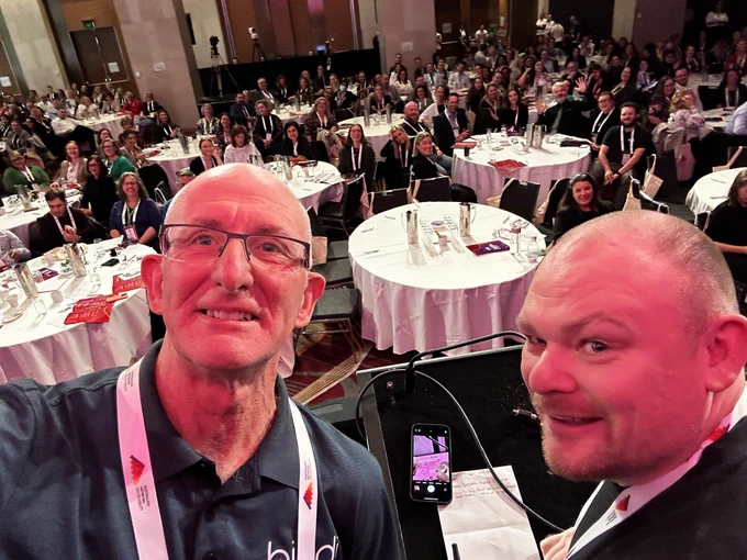 A selfie taken from the stage of the Australian Network on Disability conference in Sydney. On the left is Tony Burrett and on the right is Michael Perry.  The background is the conference crowd seated at round tables.