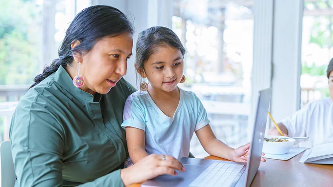 Photo of an Indigenous woman sitting with her child on her lap. They are both staring at a laptop on a table in front of them.