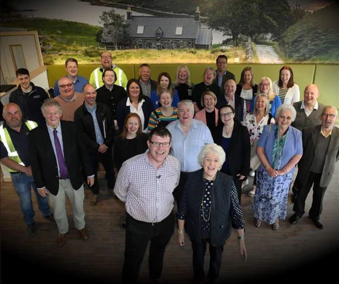 Photo of a group of people in front of a photographic mural which shows a stone house in a Highland landscape. The mural is on the wall of the cafe in the brand new Haven Centre in Inverness.