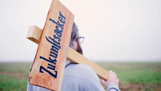 Eine Person steht auf einem Feld und trägt ein Holzschild über der Schulter mit der Aufschrift: Zukunftsacker.