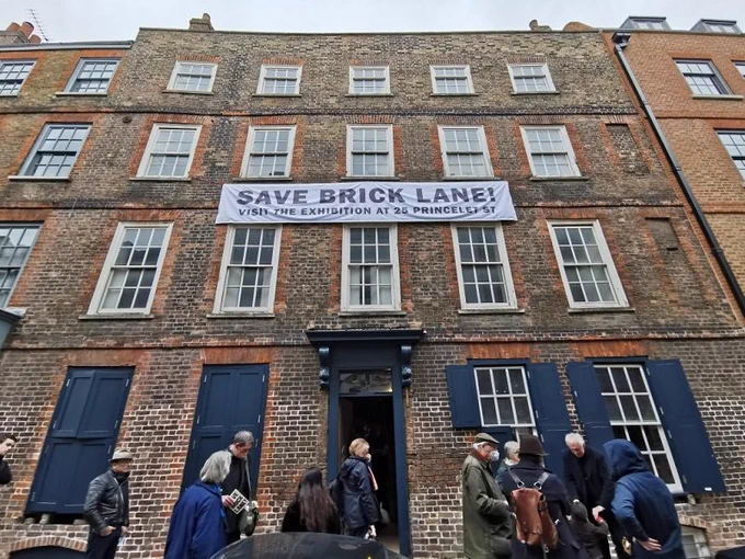 People are gathered outside 25 Princelet Street in London, waiting to see an exhibition inside the building. There is a banner on the building, which reads 'Save Brick Lane! Visit the exhibition at 25 Princelet Street.' 
