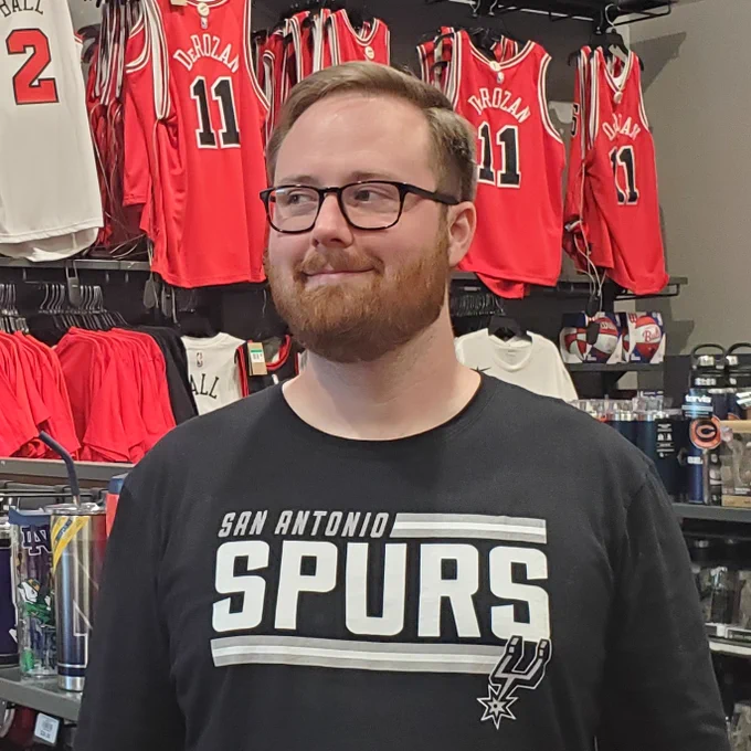 Me wearing a San Antonio Spurs shirt (my hometown team) in front of several Chicago Bulls jerseys and other Chicago sports paraphernalia at a sporting goods store. The jerseys primarily belong to DeMar DeRozan, who was traded to Chicago from San Antonio, beginning an upheaval of the roster that will in all likelihood lead to the Spurs drafting Wembanyama.