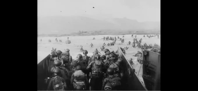 A black and white fuzzy single frame from a combat film, taken off the shore of Normandy, France - June 6, 1944.  The camera looks out at a distant shoreline fogged with gunpowder smoke, allied infantry of approximately 70 men in the frame are seen stretched all the way to the beach  wading the waist high water with their backs to the camera. In the center of the frame, the camera is perched above a landing craft, bobbing in the water with the unloading ramp already dropped down in the water. These men are about to face certain death of enemy guns entrenched ahead, with an advantage tucked into pill boxes high on the hill and shooting down at these men, as the D-Day landing commences and the start of the liberation of France begins.  Ultimately German forces are defeated in the months ahead.