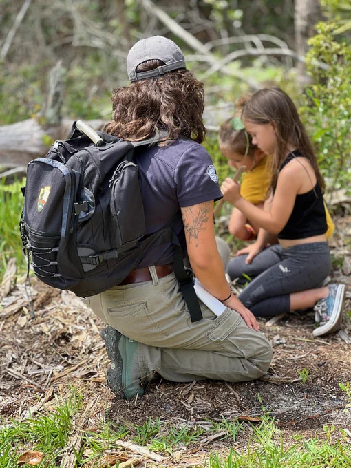 Volunteer with arrowheads on backpack kneels on ground with kids