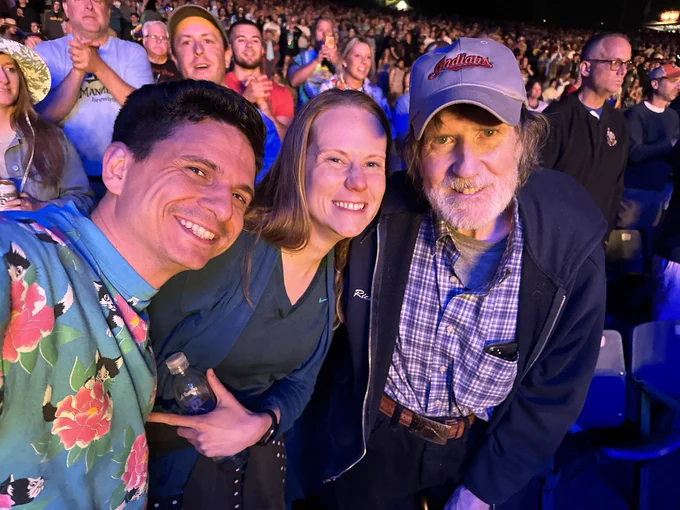 Selfie of three people in a large audience at nighttime. On the left is an attractive 30-something man with brown hair smiling and wearing a blue sweatshirt with cats and flowers on it. I, a 30-something femme person with long blonde hair am in the middle leaning towards the third person, an old man with a short white beard bending forwards slightly and smiling looking at the camera.