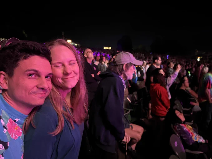Selfie of Nick and me with my dad in the background at the concert in the audience. Our skin is glowing slightly pink from the stage lights. Nick’s chin is on my shoulder and my eyes are closed. We are both smiling slightly while my dad looks forward at the out-of-image stage.