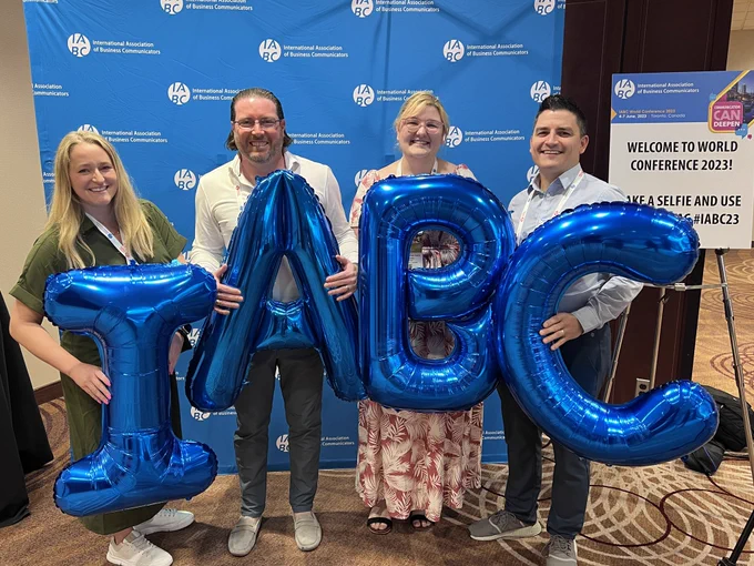 Tara, Adam, Matisse and Jason standing in front of a blue step-and-repeat banner with white IABC logos. Each holds up a blue balloon letter to spell IABC.”