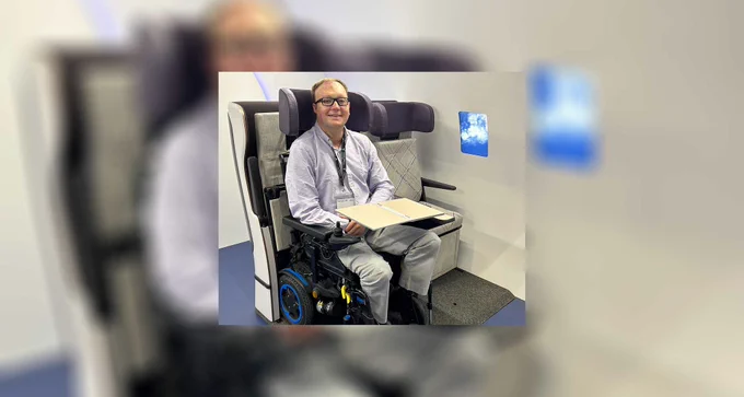 John seated in a power wheelchair in the wheelchair securement space for airplanes, with a tray table folded out across his lap.