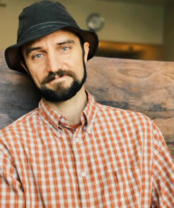 Professor Kevin Coleman wearing a hat and a checked shirt sits on a rustic bench. 