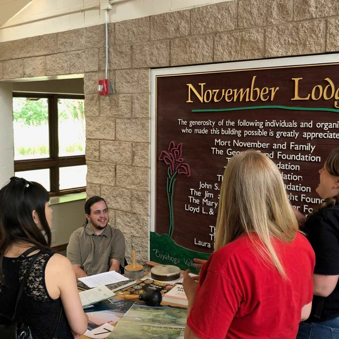 A photo of three standing people talking to someone seated at a table covered with display items. 