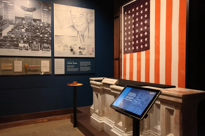 Photo of exhibit at the Sam Rayburn Museum with the white marble Speaker's Rostrum, with flag behind and touchscreen in front.