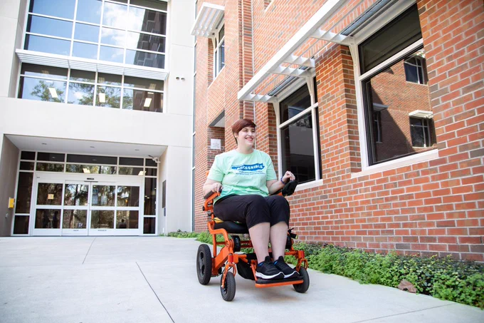 University of Florida student, Kaeli, posing in front of Cypress Hall on campus. Kaeli is looking away from the camera and smiling. She is wearing a mint green t-shirt with blue wording on it and black pants. There is one orange and one blue bracelet on her wrist. 