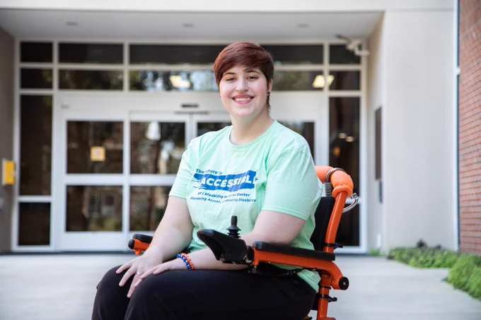 University of Florida student, Kaeli, posing in front of Cypress Hall on campus. Kaeli is smiling directly at camera with her hands in her lap. She is wearing a mint green t-shirt with blue wording on it and black pants. There is one orange and one blue bracelet on her wrist. 
