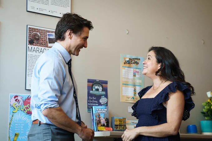 Prime Minister Justin Trudeau is standing indoors and speaking with a woman. Both of them are smiling. Posters are visible on the wall behind them.