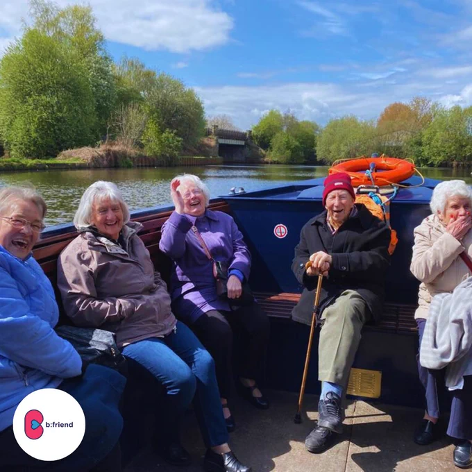 5 older neighbours sitting on a barge, laughing. Behind them is the river, blue skies and greenery. 