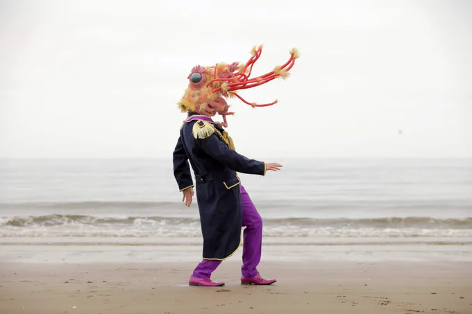 A performer wearing the party shrimp head (an oversized, hyper-realistic, fabric shrimp head) and a decorated military jacket dancing alone on a beach, they face the right and the sea is hazy and pale, blending in to the sky