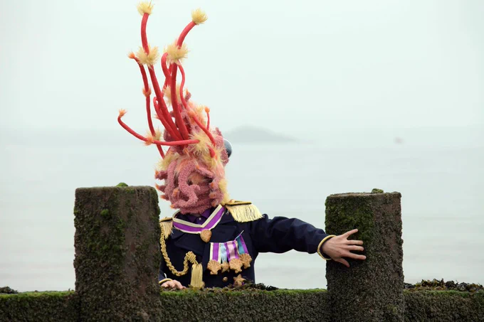A performer wearing the party shrimp head (an oversized, hyper-realistic, fabric shrimp head) and a decorated military jacket peering over a weathered stone wall, the sea is hazy and pale behind them