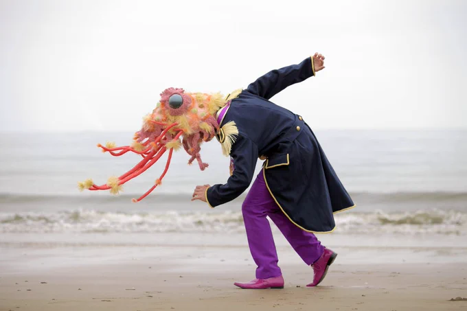 A performer wearing the party shrimp head (an oversized, hyper-realistic, fabric shrimp head) and a decorated military jacket dancing alone on a beach, they face the left and are leaning down and the sea is hazy and pale, blending in to the sky