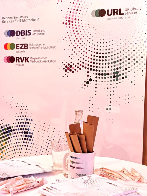Foto zum Community-Treffen des URL - UR Library Services zeigt einen Tisch mit Give-aways (Blöcke, Tasse mit Stiften, Flyern und Süßigkeiten) vor einem URL-Poster "Kennen Sie unsere Services für Bibliotheken? DBIS - Datenbank Infosystem, EZB - Elektronische Zeitschriftenbibliothek und RVK - Regensburger Verbundklassifikation"