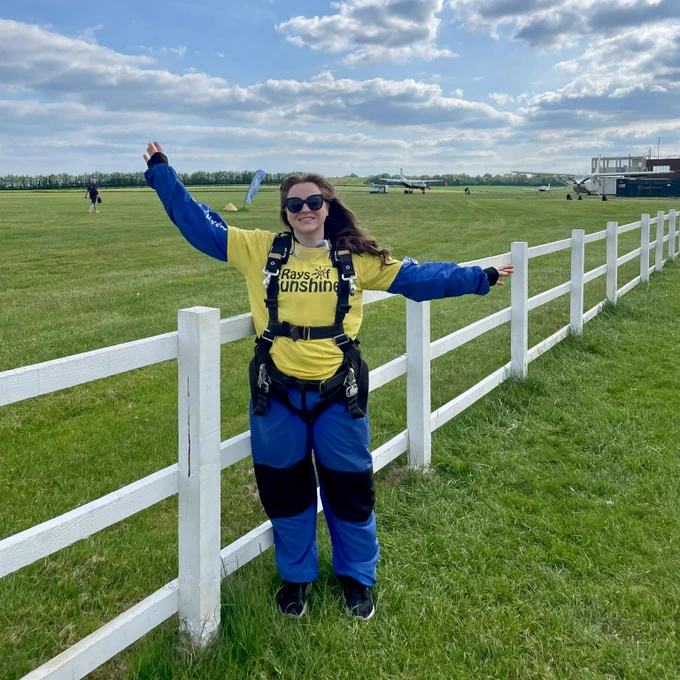 A female in blue and yellow skydiving gear. Her hands are high and she is wearing sunglasses.