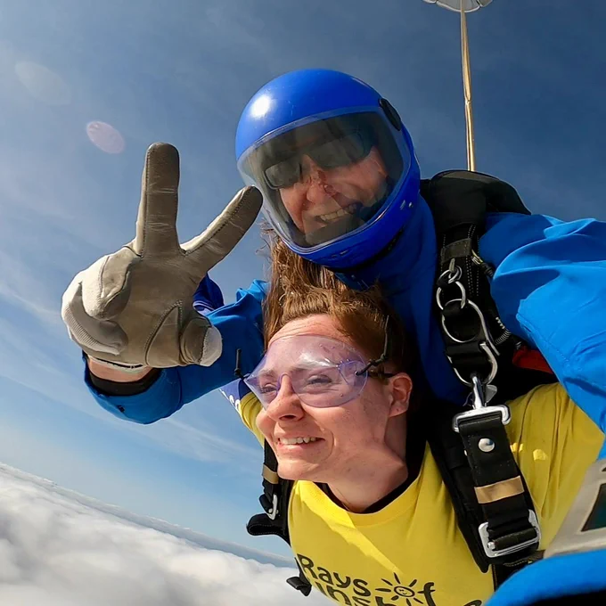A white female skydiving with an instructor fastened on her. The female is in a yellow Rays of Sunshine t-shirt. The instructor is in blue skydiving gear. The instructor is doing a peace sign to the camera.