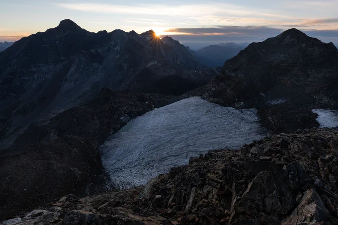 A close-up photograph of a mountain with the sun behind a peak.