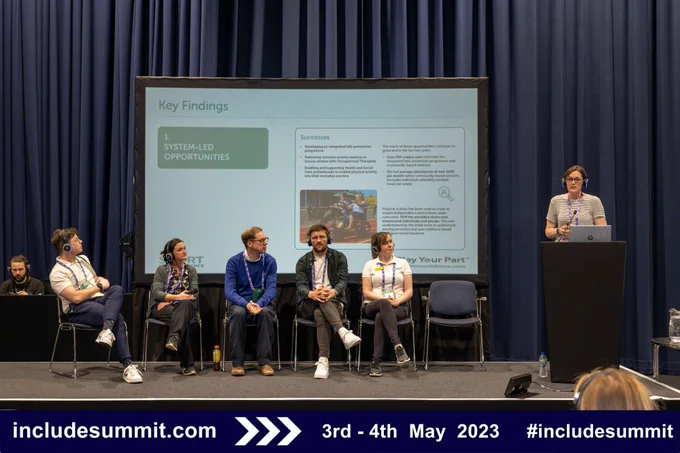 A group of five people sit on a stage inside a conference centre, another person stands at a lecturn speaking into a microphone. A large screen behind them displays a presentation about hardwiring physical activity into health and social care.