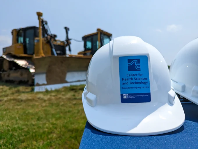 A construction hat with a label sits in the foreground, the label reads "Center fort Health and Technology Groundbreaking May 25, 2023". Bulldozers are in the background.