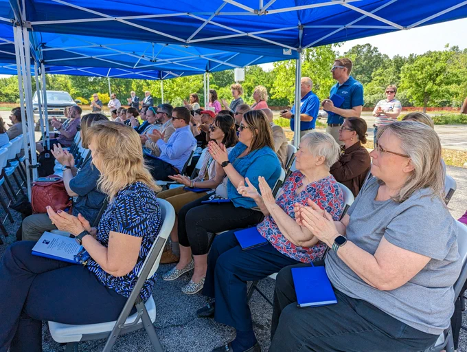 A crowd of people sit in the shade and clap at the groundbreaking ceremony. 