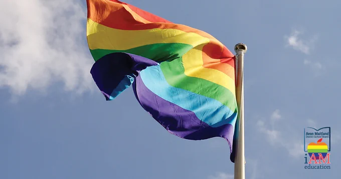 Photo of traditional rainbow Pride flag flying against blue sky. Rainbow AMDSB logo. 