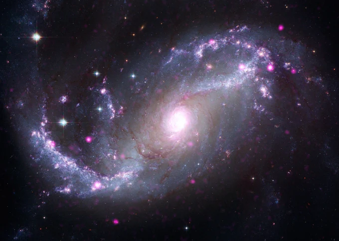 An image of spiral galaxy NGC 1672. Shown face on, this spiral galaxy has two major arms curving away from the bright swirling light at its core. One arm extends to our lower left with a gentle upward curve. The other extends to our upper right with a curve reminiscent of a question mark. Both arms have a cloudy, silver blue quality, and are dotted with bright white and purple stars of varying sizes. In this galaxy, categorized as a "barred" spiral, the arms don’t appear to reach the bright core. The space around the bright pinkish core is a swirl of murky, pale silver cloud.