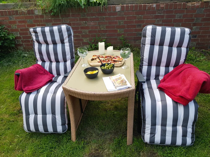 Two reclining garden chairs with grey and white striped seat covers, either side of a garden table with food and drink