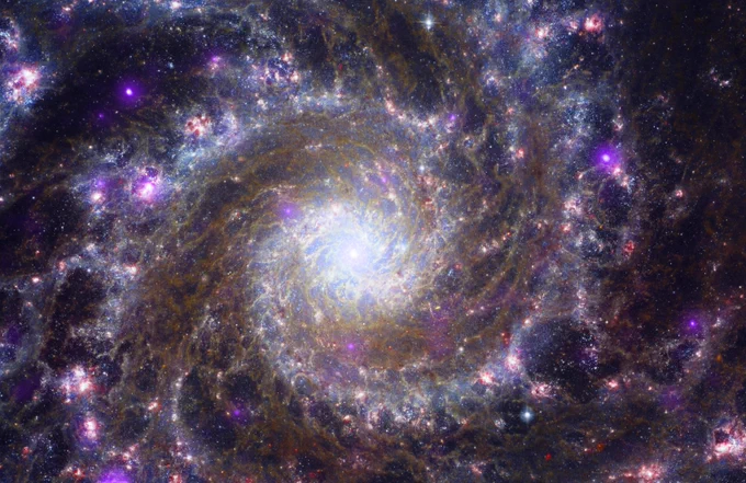 This is an image of a spiral galaxy like our own Milky Way, known as Messier 74. Shown face-on from our vantage point on Earth, the galaxy's sparkling arms spiral out from a bright white core. The core appears vibrant and alive, and crackles with lightning-like, pale blue light. Glowing, high-energy stars in purple, white, and orange, dot the lengths of the spiraling arms. Webs of murky dust crisscross the space between the curving silver blue arms, also known as dust lanes.