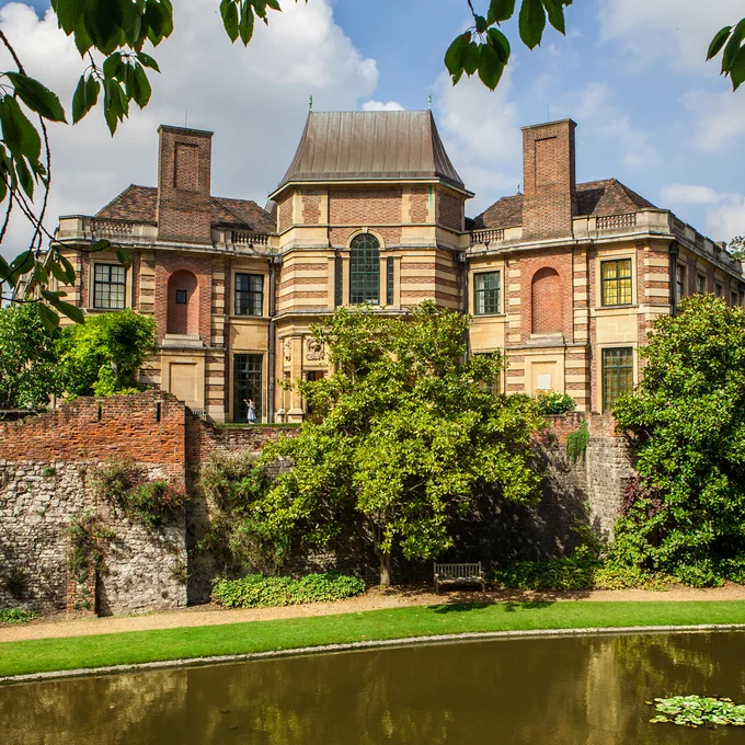 An image of an art deco palace constructed from layers of brick and beige stone. The building  overlooks a pretty garden path that features a large pond and a shaded seating area.