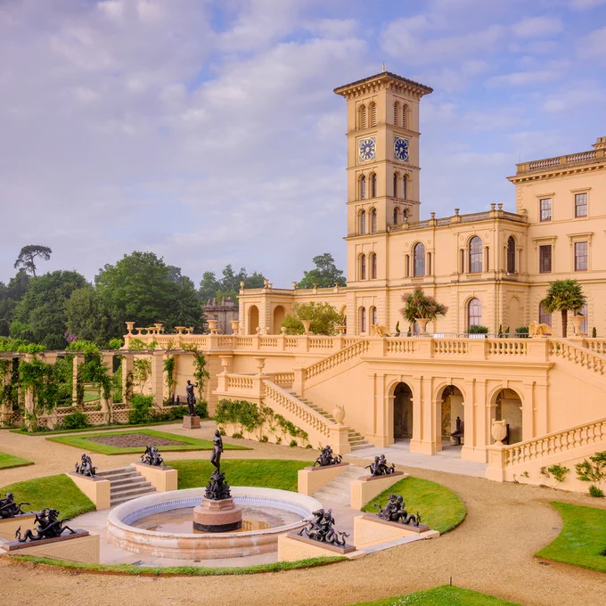 An image of an elaborate cream-coloured palace with a tall clock tower in the centre. The building sits behind neat gardens which feature a circular fountain and a collection of black statues. 