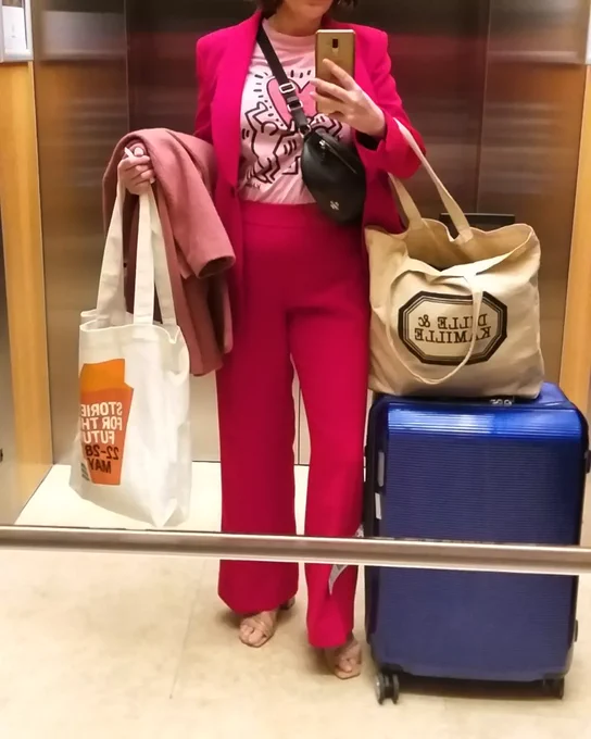 A lift mirror selfie of a woman wearing a bright red suit, a pink Keith Haring t-shirt, a black bumbag, and heels. She had luggage and tote bags with her in the lift.