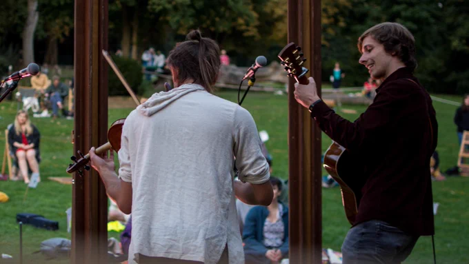 Two musicians are playing music on a violin and a guitar to a crowd of people in a sunny park.