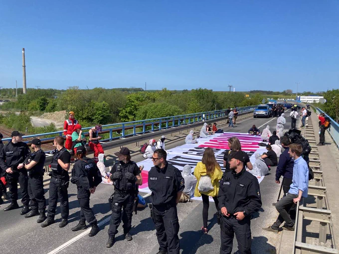 Sitting blockade with banner (caption 'Exit Gas Decolonize Now!) Police in front,riot medics ready in the background