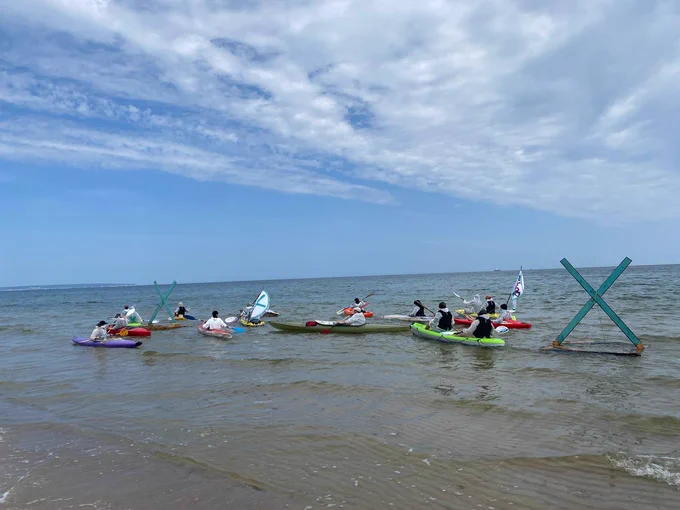 Activists in kajaks in the sea, next to a turquoise X in the sand