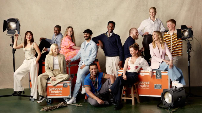A shot of 12 people under the age of 30. The photograph is taken in a photography studio. With a beige backdrop curtain and black floor. There are three large orange electrical equipment trunks that have the National Theatre logo on in white. The young people sit on, lean against and stand around the trunks at different levels. They all wear smart casual clothing. On either end there is a large spotlight at head height that two different young people lean on.