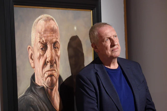 Artist Peter Howson stands in front of a painted self-portrait. Photography Greg Macvean.