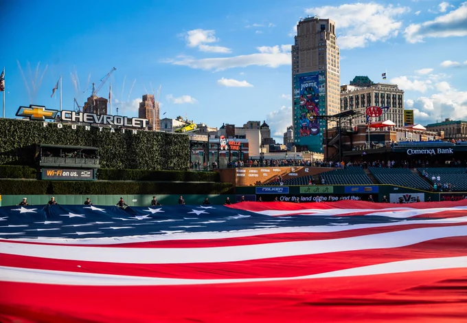 Field level view of the United States flag outstretched across the Comerica Park outfield, facing toward right field and the downtown Detroit skyline