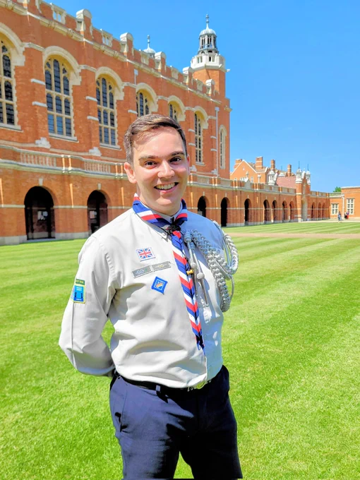 Jake in scout uniform, stood on a lawn beside the school chapel, built in red brick set against a deep blue sky.