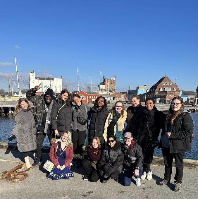 A group of people stand near a dock as they pose for a photo. Buildings and water can be seen in the background.