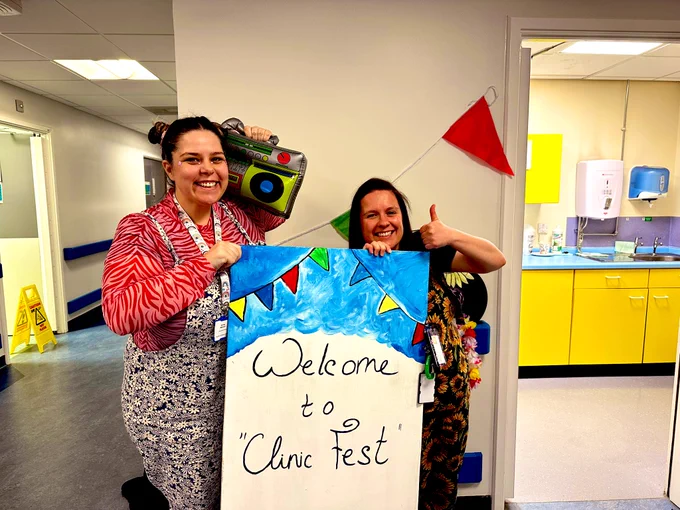 2 women holding a board say “welcome to clinic fest” wear colourful dungarees, face gems, smiling - one is holding a inflatable ghetto blaster an the other has her thumb up. Bunting in the background