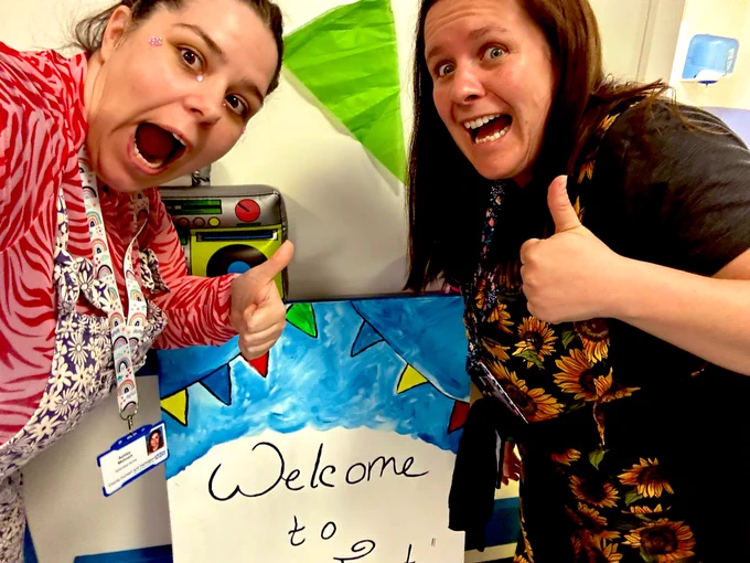 2 women stood with a board saying “welcome to clinic fest” wearing colourful dungarees, face gems, doing OMG face - with  thumbs up. Bunting in the background